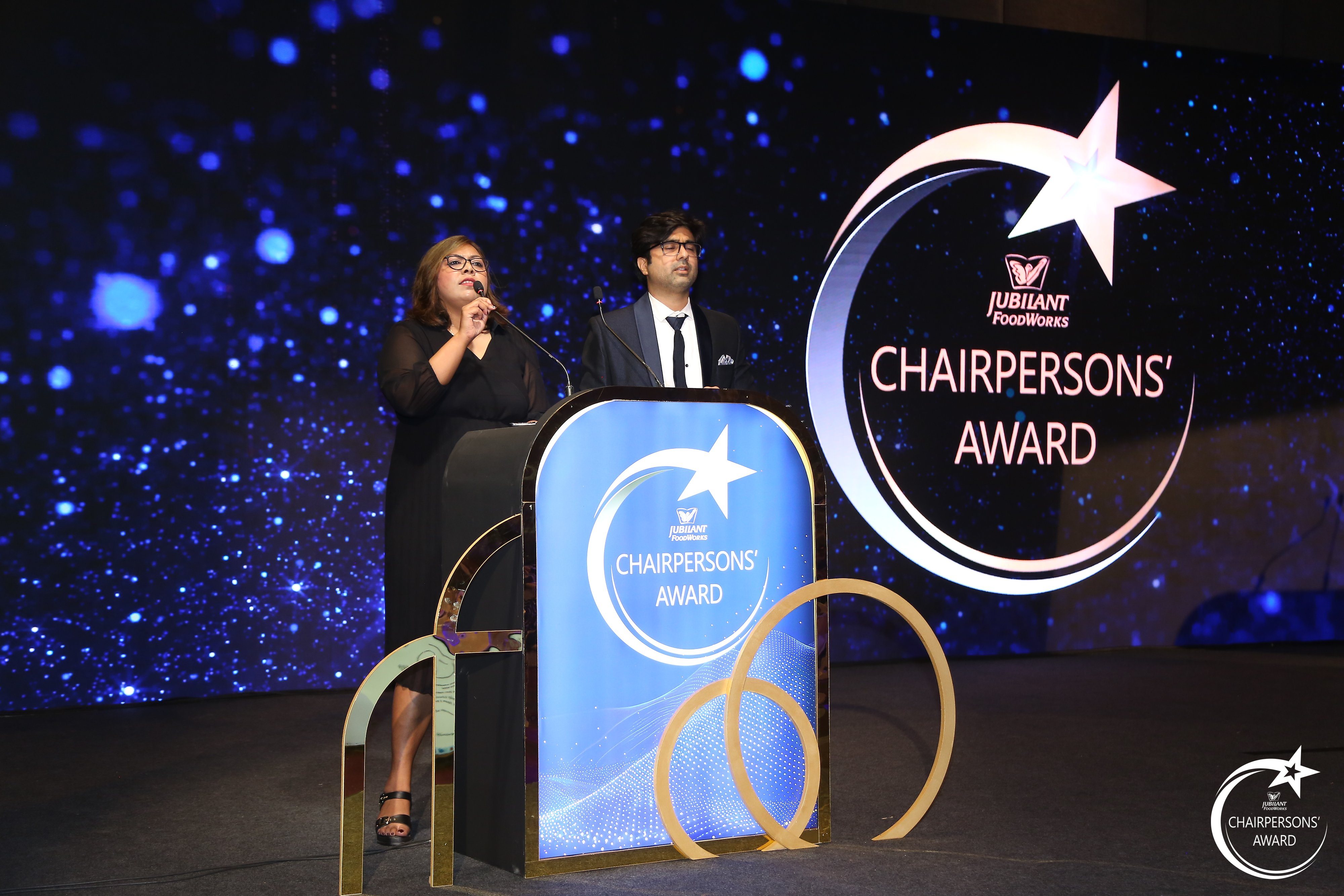Hosts on stage at the Jubilant FoodWorks Annual Chairpersons' Award beside the illuminated award crest