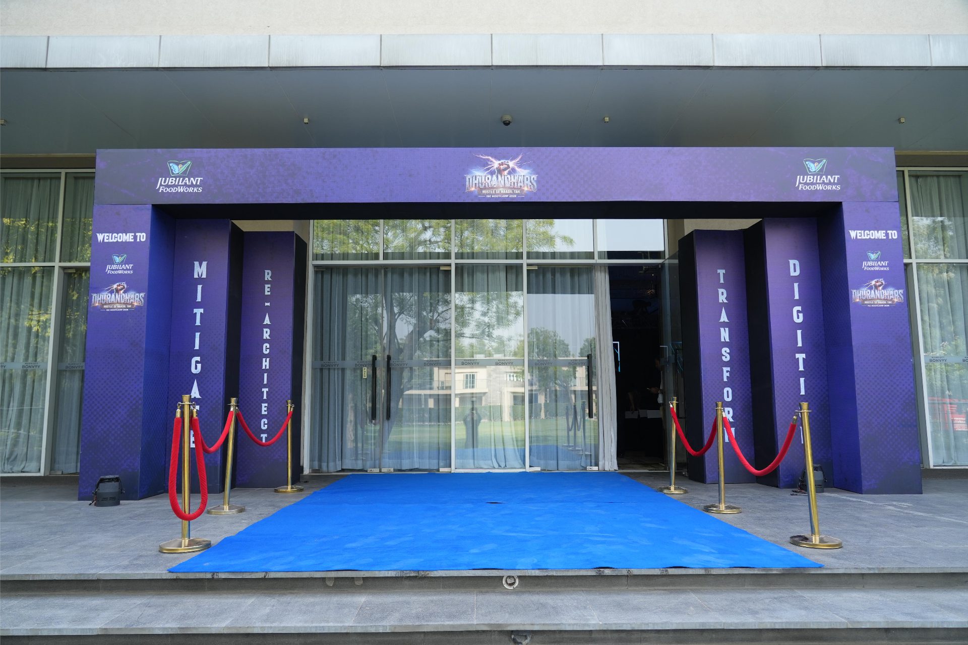 Blue-carpet entry through the Dhurandhars welcome portal, flanked by pillars reading Mitigate, Re-Architect, Transform and Digitise